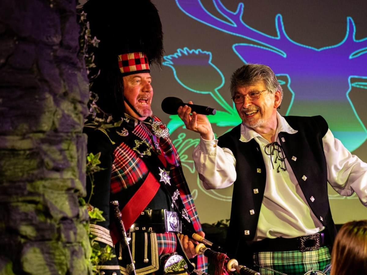 Two men in Scottish attire on stage, one with a microphone.