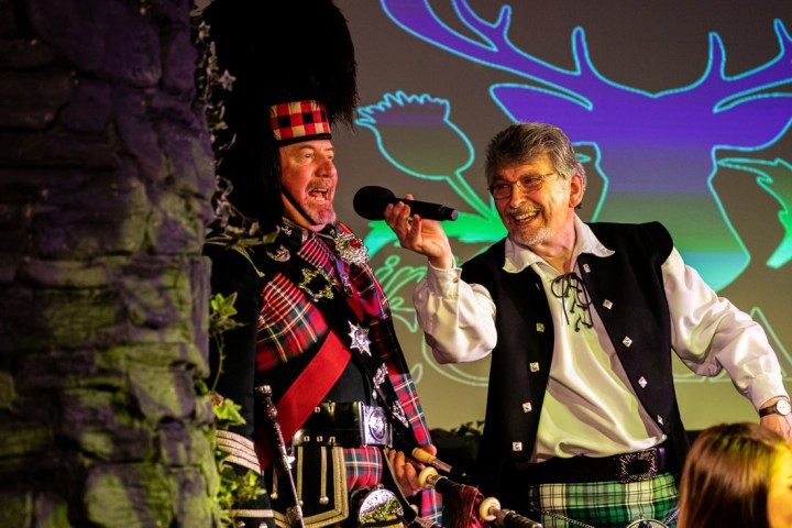 Two men in Scottish attire on stage, one with a microphone.