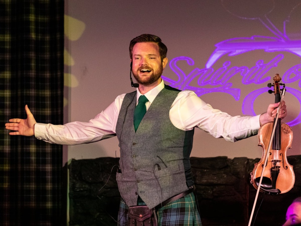 Man in Scottish attire holding a violin, arms outstretched on stage.