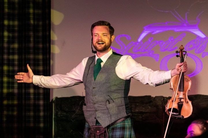 Man in Scottish attire holding a violin, arms outstretched on stage.