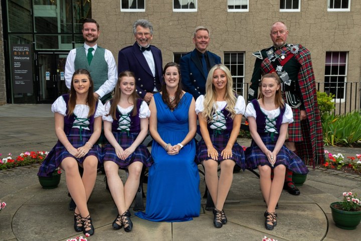 Group photo of six women in traditional attire and three men in kilts outdoors.