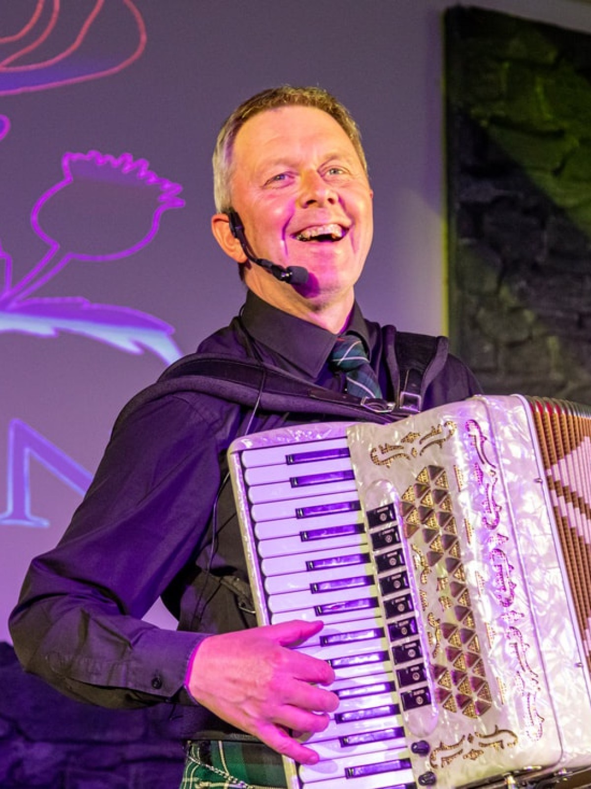 Smiling man playing accordion on stage with purple-lit background.