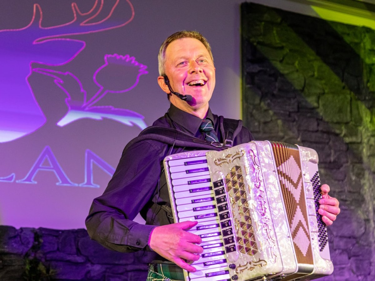 Smiling man playing accordion on stage with purple-lit background.