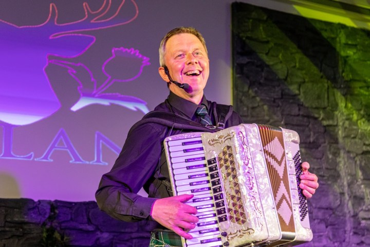 Smiling man playing accordion on stage with purple-lit background.