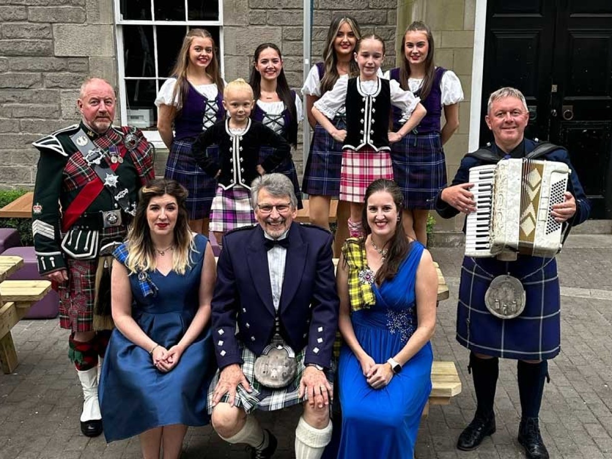 Group in traditional Scottish attire, with women in dresses and men in kilts, one playing an accordion.