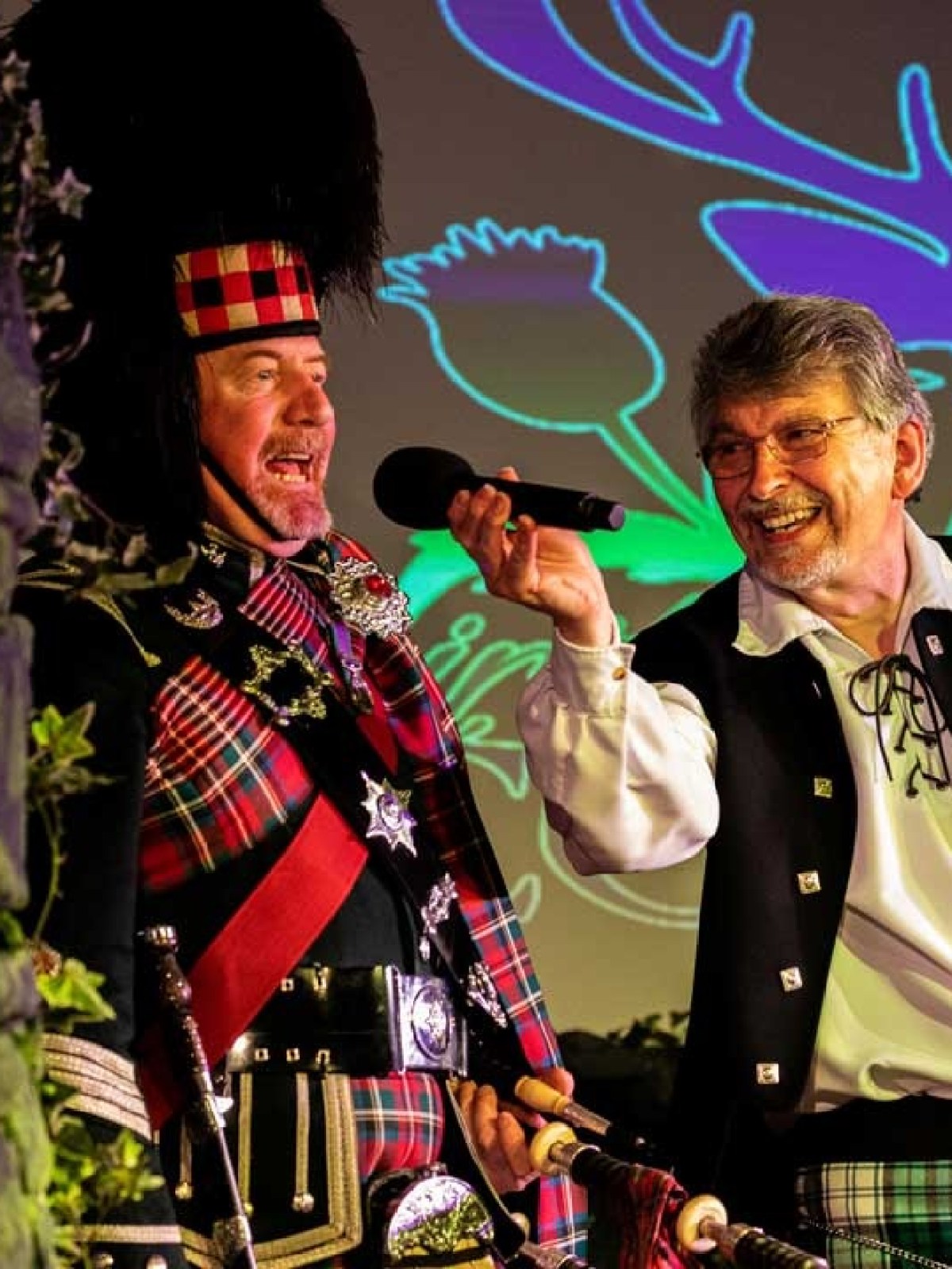 Two men in Scottish attire with microphone, backdrop silhouette of a stag.