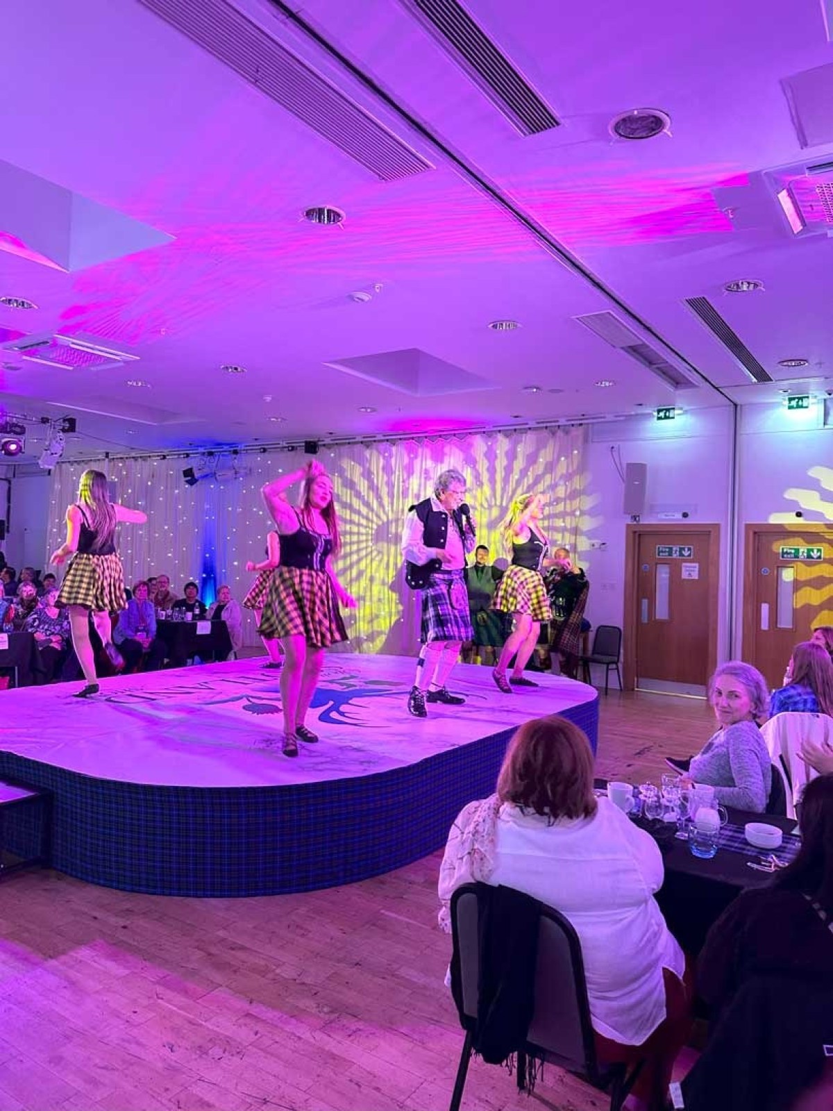 Performers in tartan skirts dance on stage with colorful lights, audience seated around.