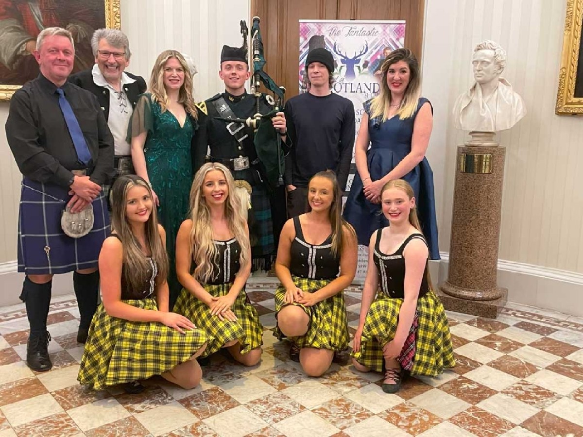 Group of people in Scottish attire, including kilts and dresses, posed indoors with a bagpiper.