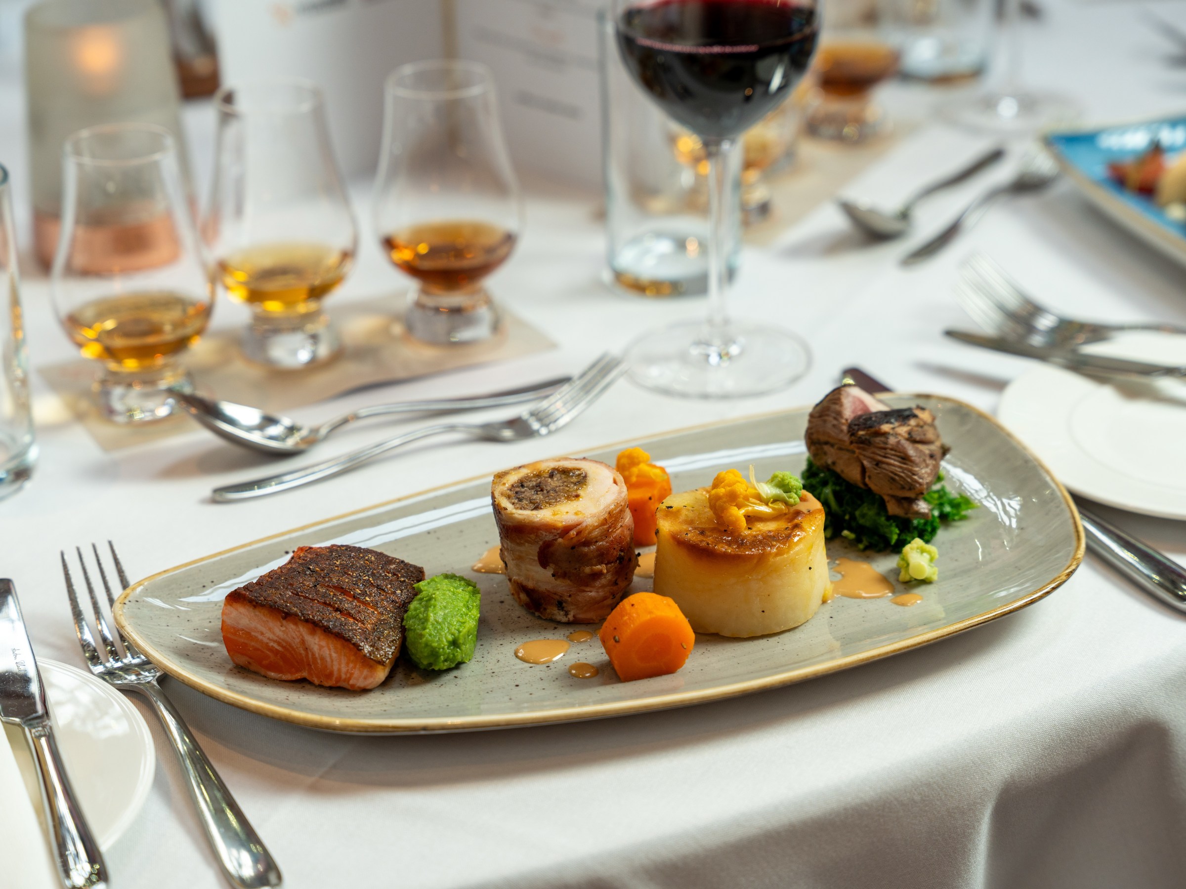 Gourmet meal with meat, salmon, vegetables, wine, and whiskey glasses on a white tablecloth.