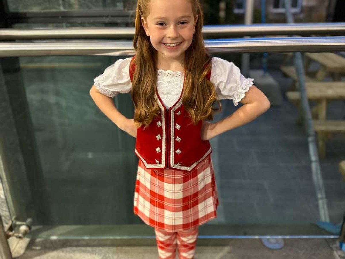 Girl in a checkered kilt outfit smiling, standing with hands on hips indoors.