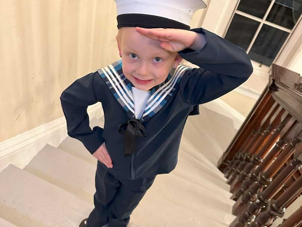 Child in sailor outfit saluting on staircase indoors.