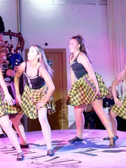 Four dancers in checkered skirts on stage with a bagpiper in the background.