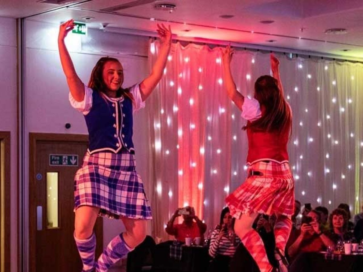 Two women in tartan skirts performing a dance, with an audience watching.