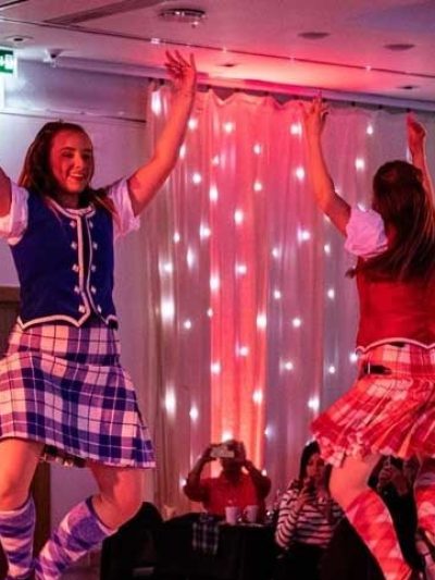 Two women in tartan skirts performing a dance, with an audience watching.