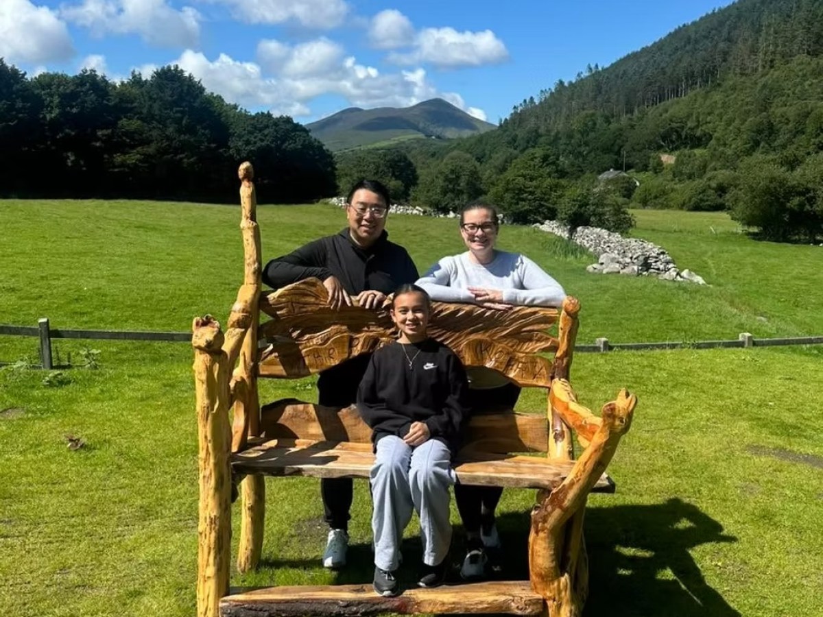Three people sitting on a large wooden bench in a grassy field with hills in the background.